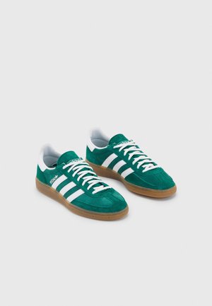 Paire de baskets en daim vert avec des bandes blanches, des lacets blancs et des semelles en caoutchouc naturel, présentées sur un fond gris clair uni.