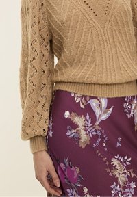 Beige neulepaita, jossa on teksturoitu ja kuvioitu design sekä pitkät hihat, yhdistettynä purppura kukkakahvilahenteeseen, jossa on erilaisia värejä ja yksityiskohtia.