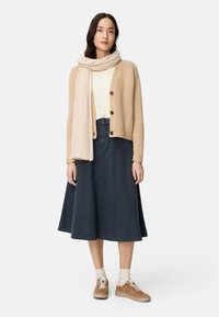 Beige gebreid vest met knopen, wit topje, donkerblauwe knielange rok en beige sneakers met witte veters en sokken.