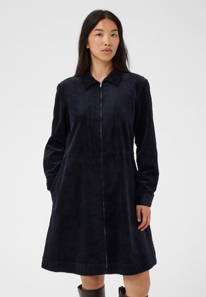 Femme portant une robe en velours côtelé bleu marine foncé à manches longues avec fermeture éclair à l'avant et col, debout devant un fond uni.