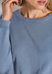 Frau in einem hellblauen Strickpullover mit geripptem Kragen und Bündchen, die sich am kurzen blonden Haar in der Nähe des Ohrs berührt.