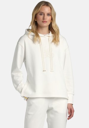 Jeune femme aux cheveux blonds portant un sweat à capuche blanc et un pantalon blanc assorti, posant avec une main dans la poche sur un fond uni.