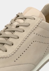 Μπεζ σουέντ αθλητικό παπούτσι με υφή στην επάνω επιφάνεια, σκαλοπατιές στις άκρες, στρογγυλά κορδόνια, διάτρητες λεπτομέρειες και γυαλιστερό κάλυμμα στη φτέρνα.