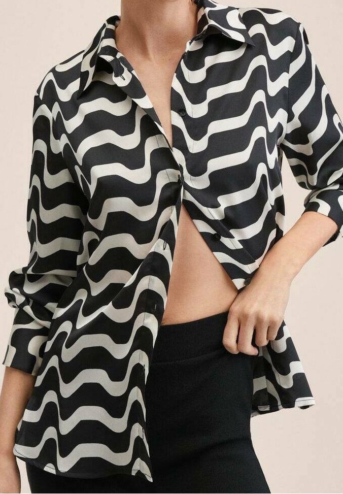 Femme portant une chemise à rayures ondulées noir et blanc, boutonnée et partiellement ouverte, associée à un pantalon noir, une main tirant légèrement la chemise sur le côté.