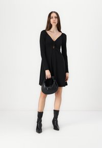 Vestido preto de manga longa com decote em V e detalhe em buraco, combinado com uma bolsa preta arredondada e botas pretas até ao joelho.