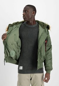 Grüne, gepolsterte Jacke mit einer mit Fell gefütterten Kapuze, Reißverschluss und Seitentaschen, getragen über einem dunklen, gestrickten Pullover mit Rippstruktur.