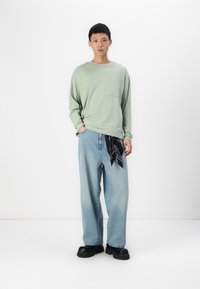 Lichtgroen overhemd met lange mouwen en een zak, gecombineerd met losse blauwe jeans, vergezeld van een zwarte gebonden bandana om de taille en zwarte chunky schoenen.