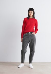 Mujer de pie con un suéter texturizado rojo, jeans grises de tiro alto con botones y zapatillas blancas frente a un fondo claro y simple.