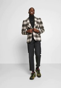 Blazer a cuadros en beige y negro sobre un cuello de tortuga negro, combinado con pantalones cargo oscuros y zapatillas con acentos verdes. Textura suave, corte ajustado.