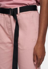 Los pantalones de algodón rosa presentan un cinturón de nailon negro, un bolsillo lateral y un detalle bordado "10" cerca del bolsillo. Textura suave y diseño ajustado.