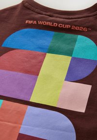 Texte FIFA World Cup 2026 au-dessus d'un design géométrique coloré sur un tissu marron, avec des formes bleues, vertes, violettes, jaunes, roses et rouges.
