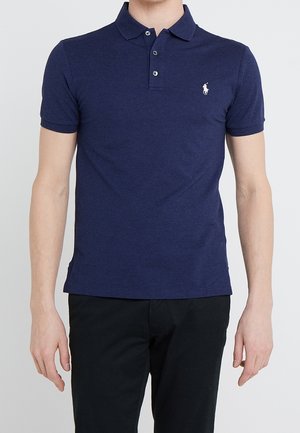 Homme portant un polo bleu marine à manches courtes avec un petit logo blanc brodé sur la poitrine gauche, associé à un pantalon noir.