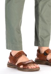 Sandales en cuir marron avec des sangles ajustables, une semelle rembourrée et un design ouvert aux orteils, associées à un pantalon vert clair.