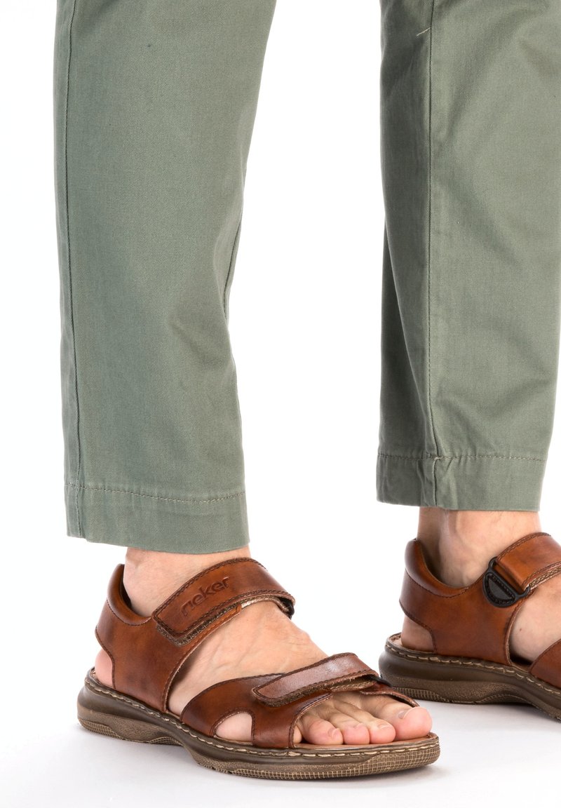 Sandales en cuir marron avec des sangles ajustables, une semelle rembourrée et un design ouvert aux orteils, associées à un pantalon vert clair.