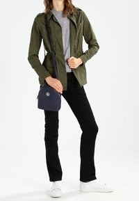 Chaqueta verde olivo con un diseño ajustado, camiseta gris, pantalones negros, zapatillas blancas y un pequeño bolso cruzado azul marino con un detalle de logo.