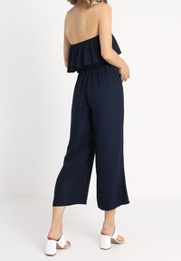 Žena oblečená v bezramínkovém námořnickém skákacím oblečení s volánkovými detaily a širokými nohavicemi, doplněná bílými sandály na blokovém podpatku, stojící na bílém pozadí.