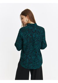 Czarna koszula z długim rękawem w turkusowy abstrakcyjny wzór liści, z charakterystycznym kołnierzykiem oraz guzikami na plecach. Miękka tekstura materiału.
