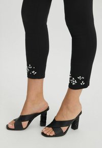 Mule negre cu toc înalt, cu un design cu bretele încrucișate și un toc bloc cu un model. Blugi negri, ajustați, cu embellismente asemănătoare cu perlele la tiv.