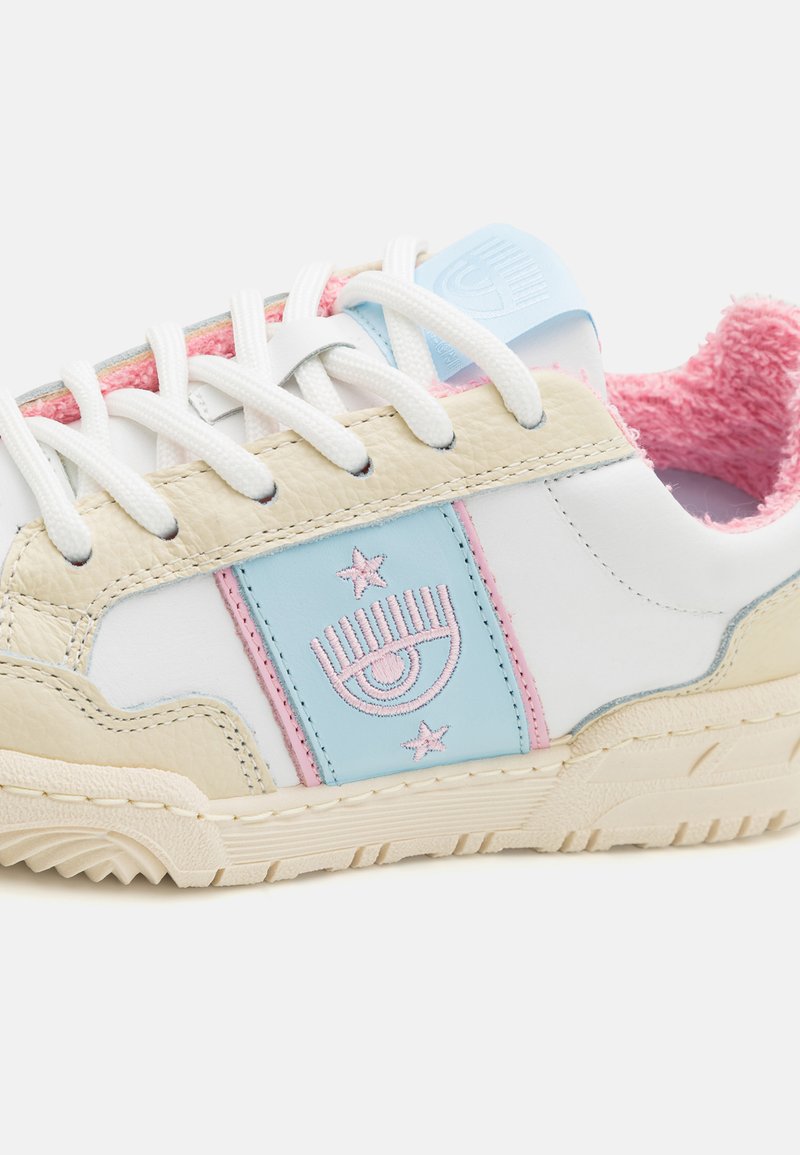 Les baskets présentent une tige en cuir blanc, des accents beiges, des éléments de design rose et bleu, ainsi que des lacets blancs avec une doublure texturée rose.