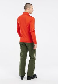 Punainen pitkähiha, jossa on korkea kaulus, yhdistettynä vihreisiin housuihin, joissa on vetoketjulliset taskut ja mustat kengät, nähtynä takaa.