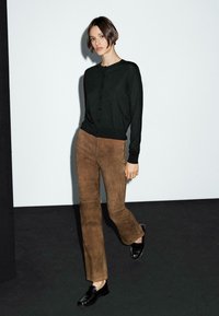 A woman walks confidently, wearing a black cardigan and brown suede pants, on a minimalistic dark floor with a light backdrop.