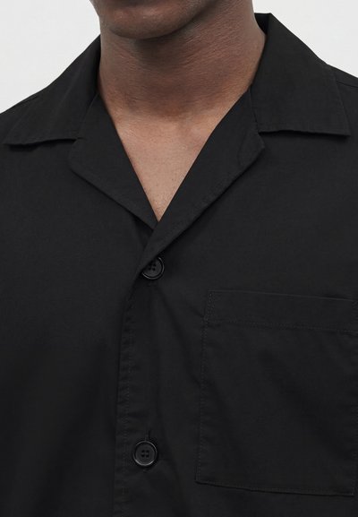 Άνδρας με μαύρο πουκάμισο με κουμπιά, γιακά τύπου camp και τσέπη στο στήθος, εμφανίζεται από τον λαιμό έως το μέσο του κορμού.