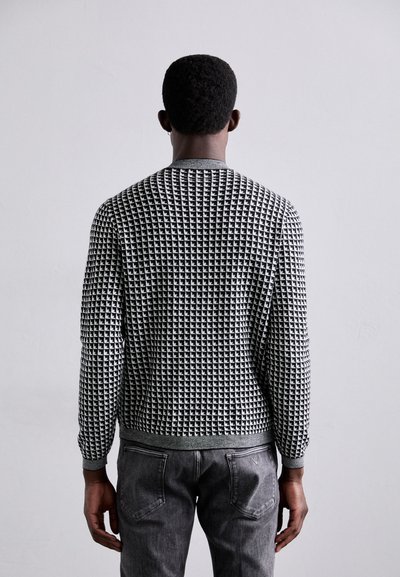 Pull à motifs noir et blanc avec un col rond et des poignets côtelés, associé à un jean gris, vu de dos sur un fond neutre.