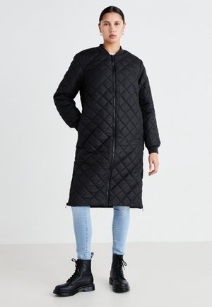 Manteau long matelassé noir avec fermeture éclair, doté de poches latérales et de poignets côtelés, associé à un jean slim bleu clair et des bottes à lacets noires.