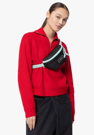 Zwarte heuptas met mint-kleurige accenten, ritssluiting, verstelbare schouderband en logo-embleem. G gedragen over een rode gebreide trui en donkere jeans.
