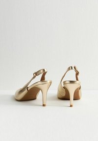 Un par de zapatos de tacón alto metálicos dorados tipo slingback con correas en el tobillo, vistos desde la parte de atrás sobre una superficie blanca.