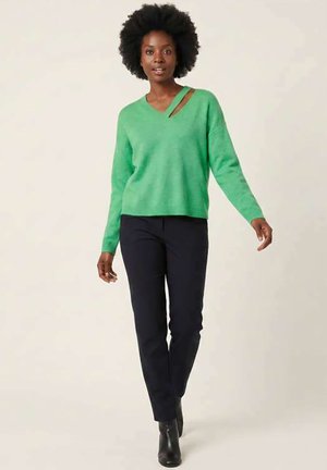 Femme marchant portant un pull vert avec une découpe à l'épaule, un pantalon noir et des bottines noires sur un fond beige uni.