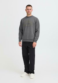 Sweatshirt gris avec un design imprimé en or et vert, col rond, manches longues, associé à un jean noir et des baskets blanches.