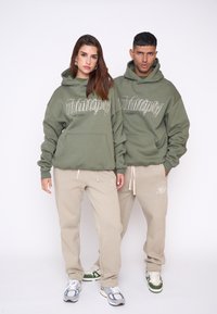 Felpe verde oliva con tasche frontali, decorate con testo bianco "multiply"; abbinate a pantaloni della tuta beige con accenti ricamati.