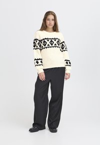 Maglione lavorato a maglia color crema con motivo geometrico nero, maniche lunghe, scollatura rotonda; abbinato a pantaloni neri larghi e scarpe nere flat.