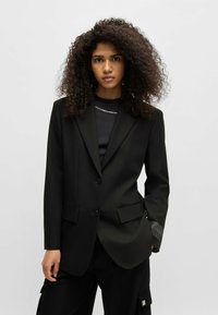 Blazer nero su misura con due bottoni, revers a disegno e tasche laterali. Dettaglio notevole: logo in rilievo sul polsino. Indossato sopra una maglia nera.