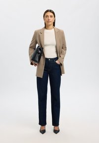 Beige blazer over a white top, dark blue jeans, black heels, and holding a black handbag with a unique design. Smooth textures, tailored fit.