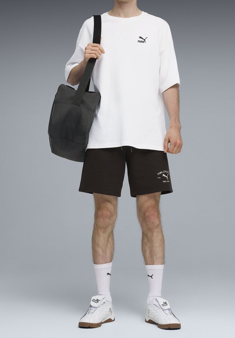 Personne portant un t-shirt blanc Puma, un short noir Puma, des chaussettes blanches Puma et des baskets blanches Puma, tenant un sac à bandoulière noir.