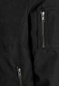 Chaqueta negra con tejido suave y texturizado; cuenta con múltiples cremalleras, incluyendo un bolsillo destacado en la manga, y costuras reforzadas.