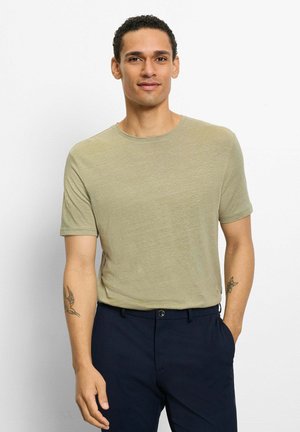 Man with short curly hair wearing a light olive t-shirt and dark navy pants, standing with one hand in pocket against a white background.