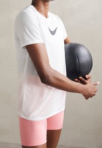 Vit idrottströja med en grå logotyp, kombinerad med rosa shorts. Personen håller en svart medicinboll som visar en slät, rund textur.