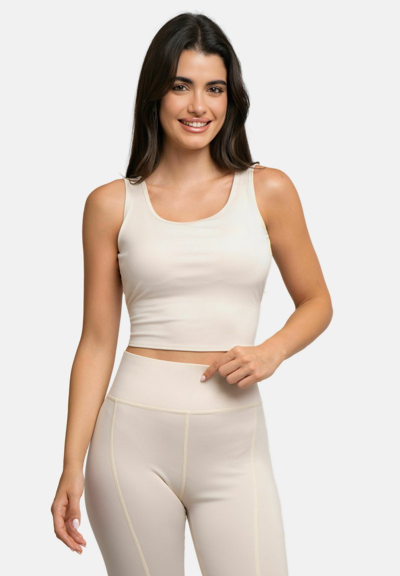 Femme aux longs cheveux foncés souriante, portant un crop top beige clair sans manches et des leggings taille haute assortis, pointant sa taille.