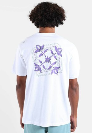 Homme aux cheveux bouclés portant un t-shirt blanc avec un motif floral violet et un texte au dos, associé à un short bleu clair.