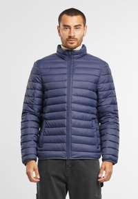Tamsiai mėlyna pūkinė striukė su aukštu apykakle, horizontaliu siūlų raštu, priekiniais užtrauktukais ir šoninėmis kišenėmis. Lengvas, elegantiškas dizainas.