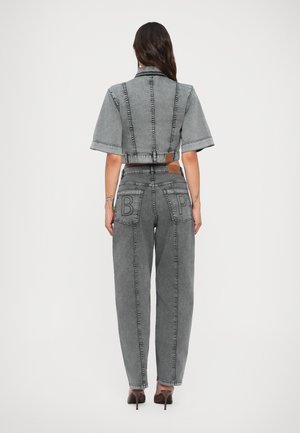 Kvinde iført grå kort denimjakke og matchende højtaljede jeans med påsyede "B" og "P" på baglommerne, stående mod en ensfarvet baggrund.