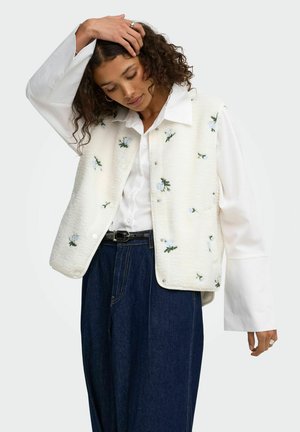 Kvinde med krøllet hår iført hvid broderet vest over hvid bluse og mørkeblå jeans med høj talje, poserer med den ene hånd på hovedet.
