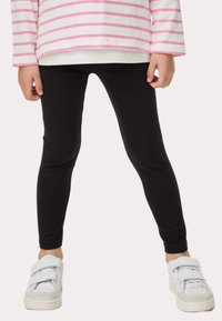 Leggings negros de tela elástica, con un ajuste ceñido. Combinados con zapatillas blancas con correas de gancho y bucle y detalles en gris.