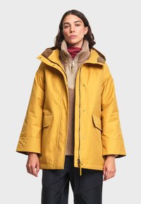 Femme portant une veste d'hiver jaune à capuche par-dessus des hauts superposés beige et bordeaux, debout devant un fond clair uni.