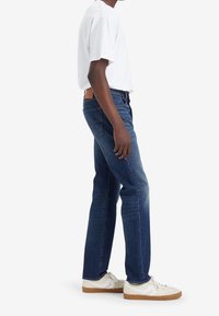 Mörkblå denimjeans med rak passform, lätt blekning och brun läderpatch på baksidan. Matchas med en vit t-shirt med kort ärm och sneakers.