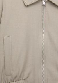Beige lynlåsjakke med broderet krave, elastisk kant og en sidelomme, lavet af et glat, tekstureret stof.