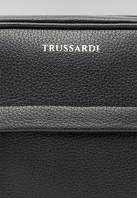 Borsa nera in pelle con superficie strutturata, logo argentato "TRUSSARDI" sul davanti e una striscia grigia al centro.